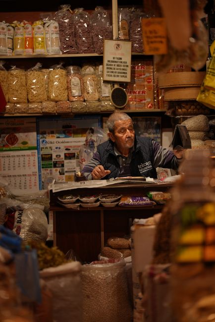 Elderly vendor tending traditional market stall with spices and grains in warm indoor setting.